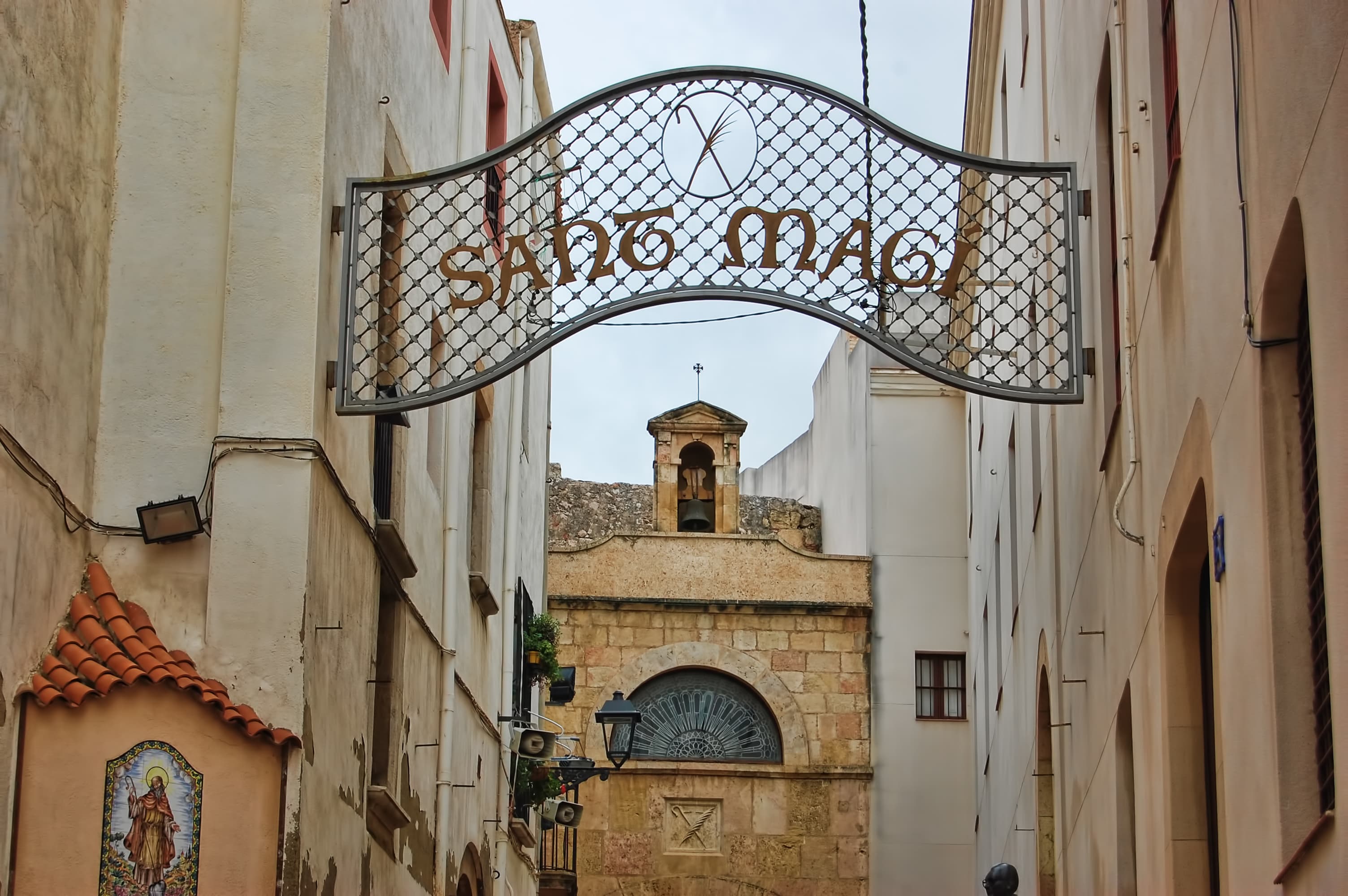 Arco de forja con el nombre «Sant Magí» sobre el callejón que lleva a la Capella de Sant Magí del Portal del Carro, en Tarragona, con el campanario al fondo