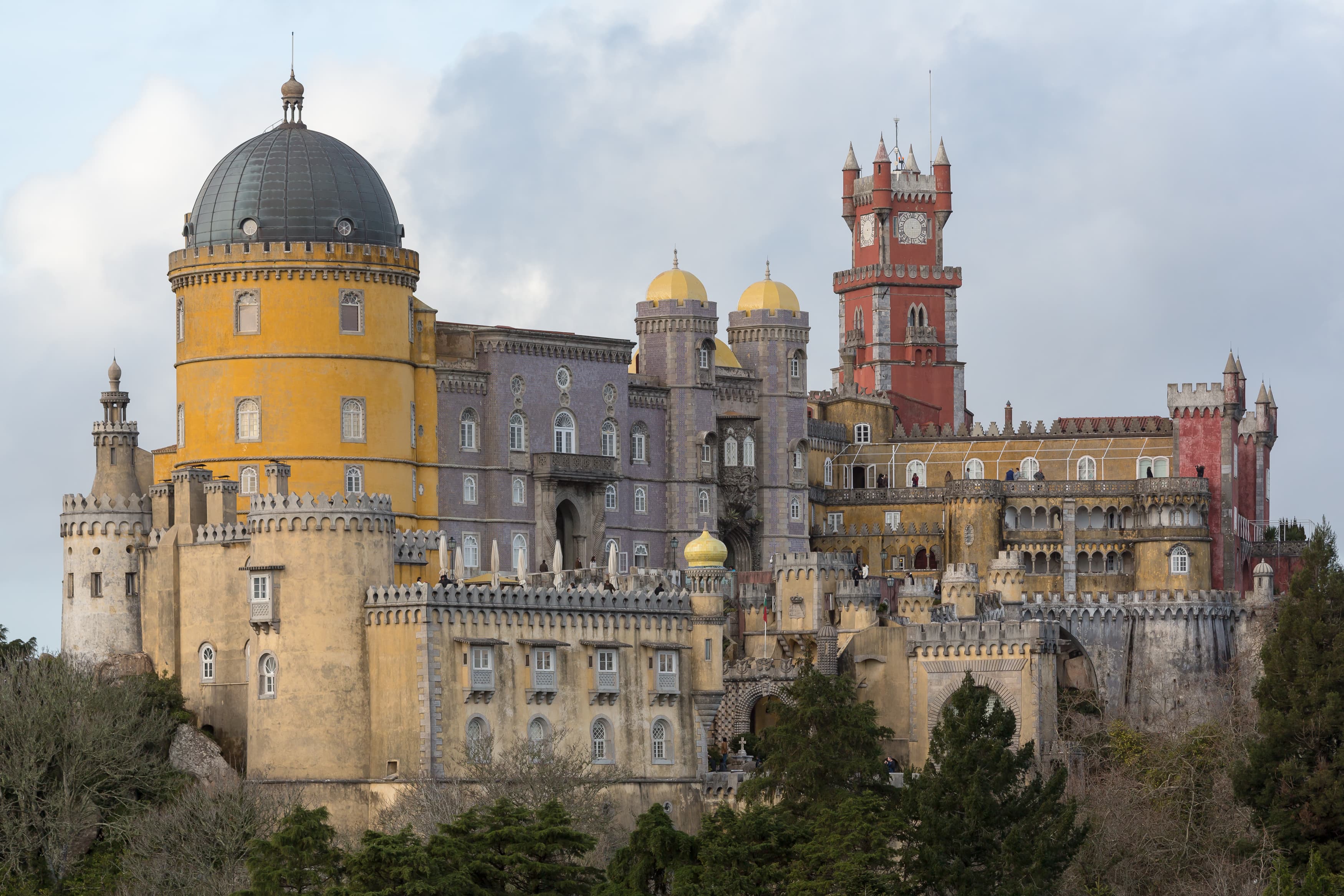 Palacio Nacional da Pena con sus torres y muros rojos y amarillos elevándose sobre la cima boscosa de la Serra de Sintra bajo un cielo despejado.